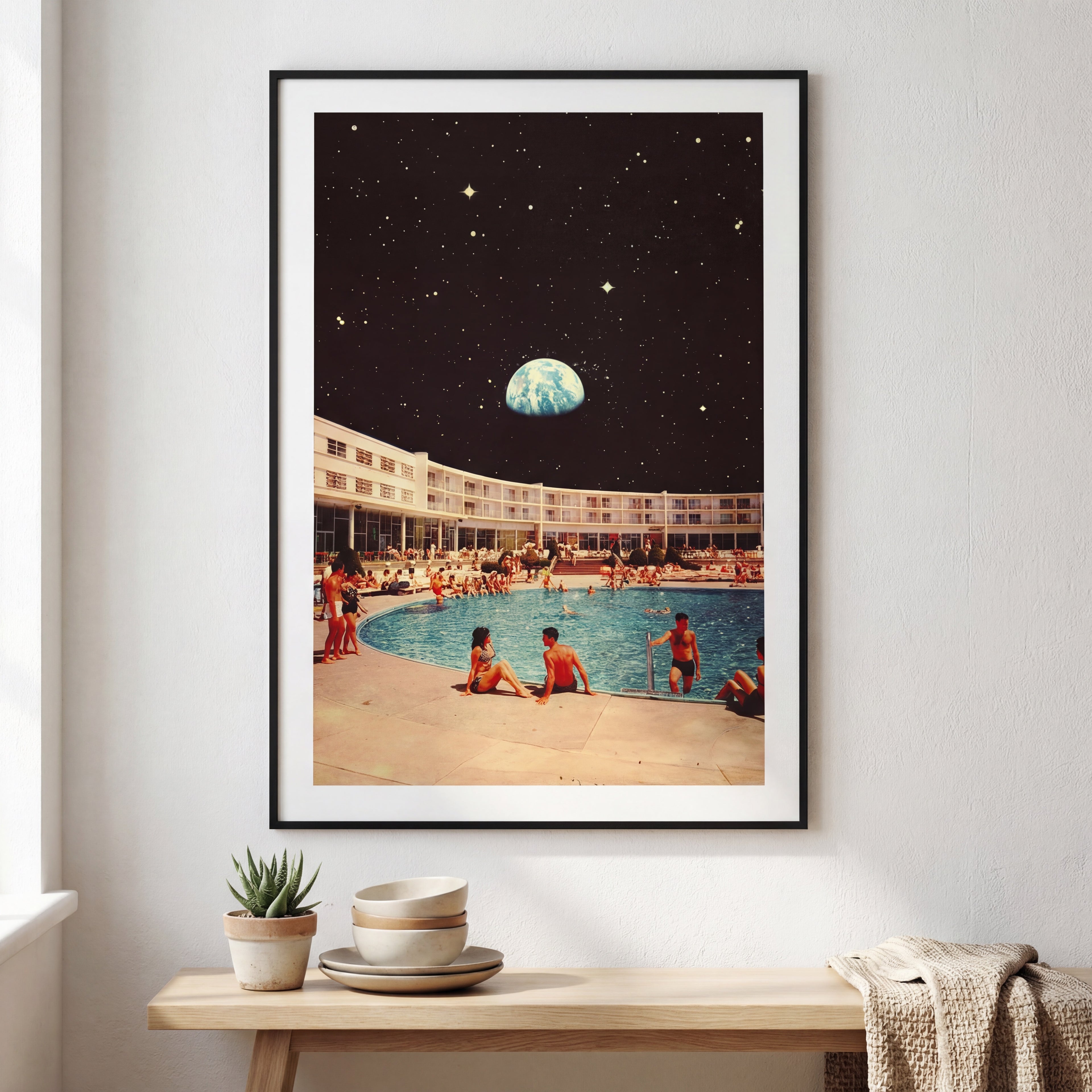 Framed artwork of a pool scene with people and a large planet-like object in the sky, displayed on a wall above a wooden table.